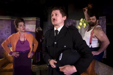 A policeman speaking to a woman with a purple top and orange cardigan. A man holdig an ornament above his head, ready to throw it.