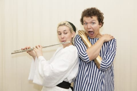 One actor in a striped tshirt with a gold prosthetic arm. The other actor in a white tshirt playing the flute.