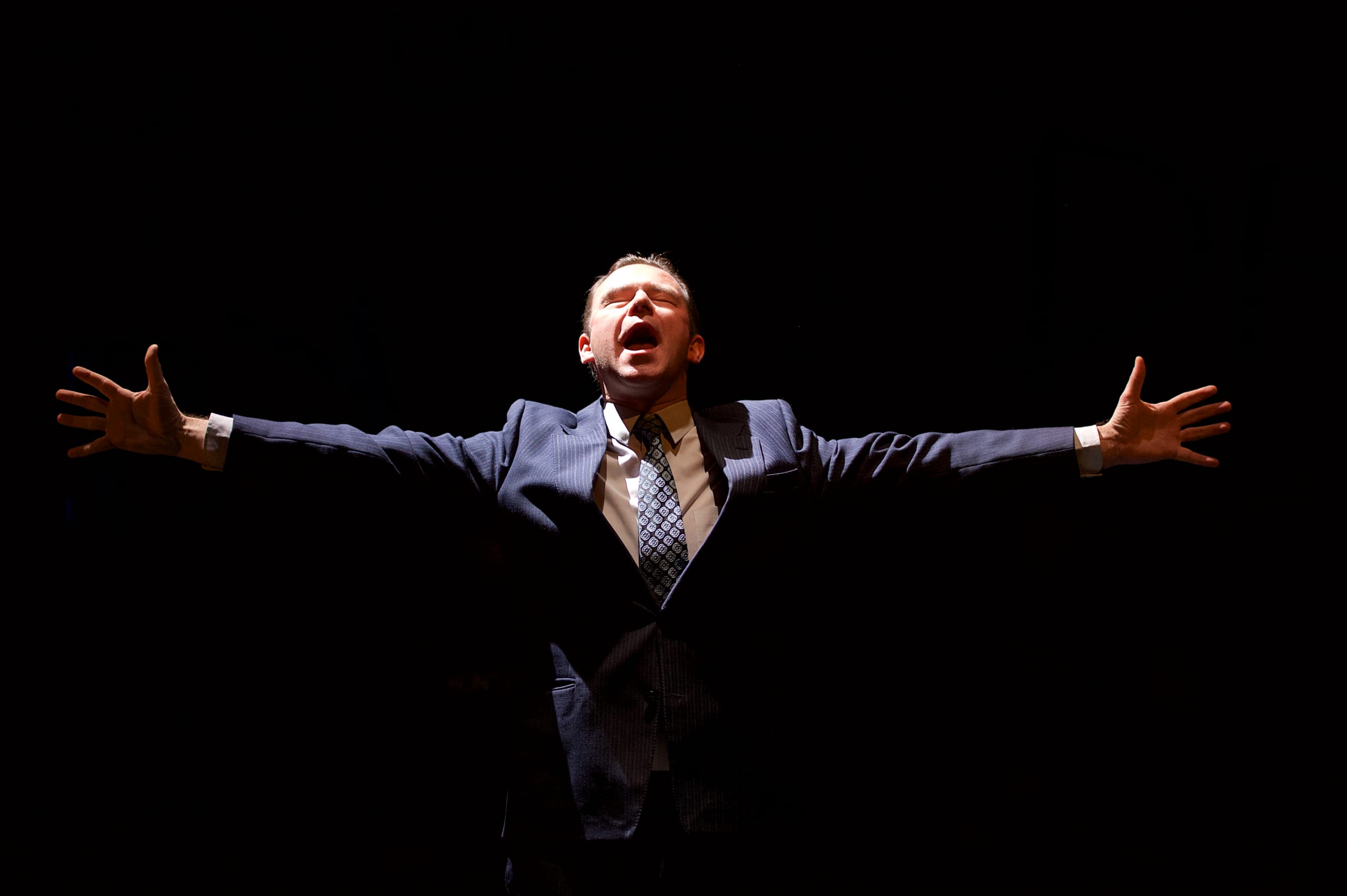 A man in a suit on dark stage with arms spread wide