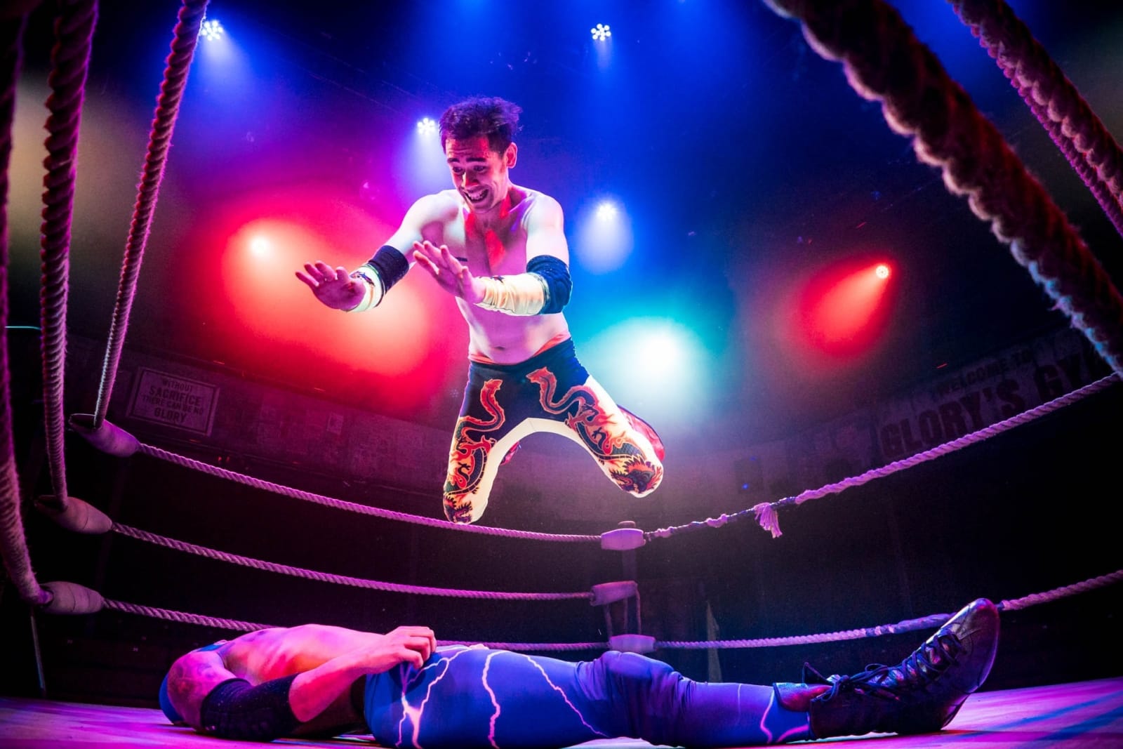 A wrestler diving onto another laid in the ring, bathed in coloured spotlights