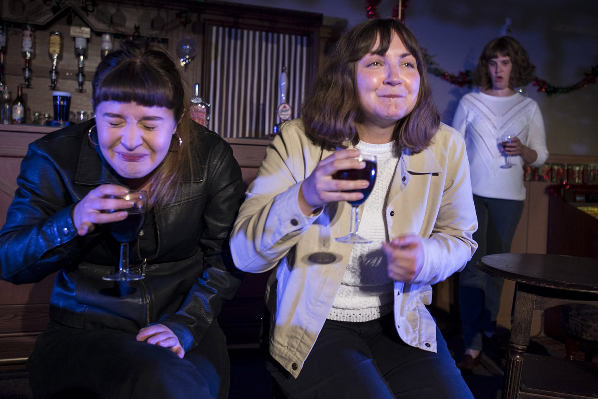 2 women in a pub, laughing. A younger version of one of the women watches them from afar.