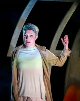 A woman in a dystopian chamber dressed in a nightgown with arms raised in defence