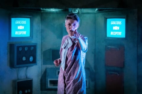 A woman in a dystopian chamber pointing to the audience