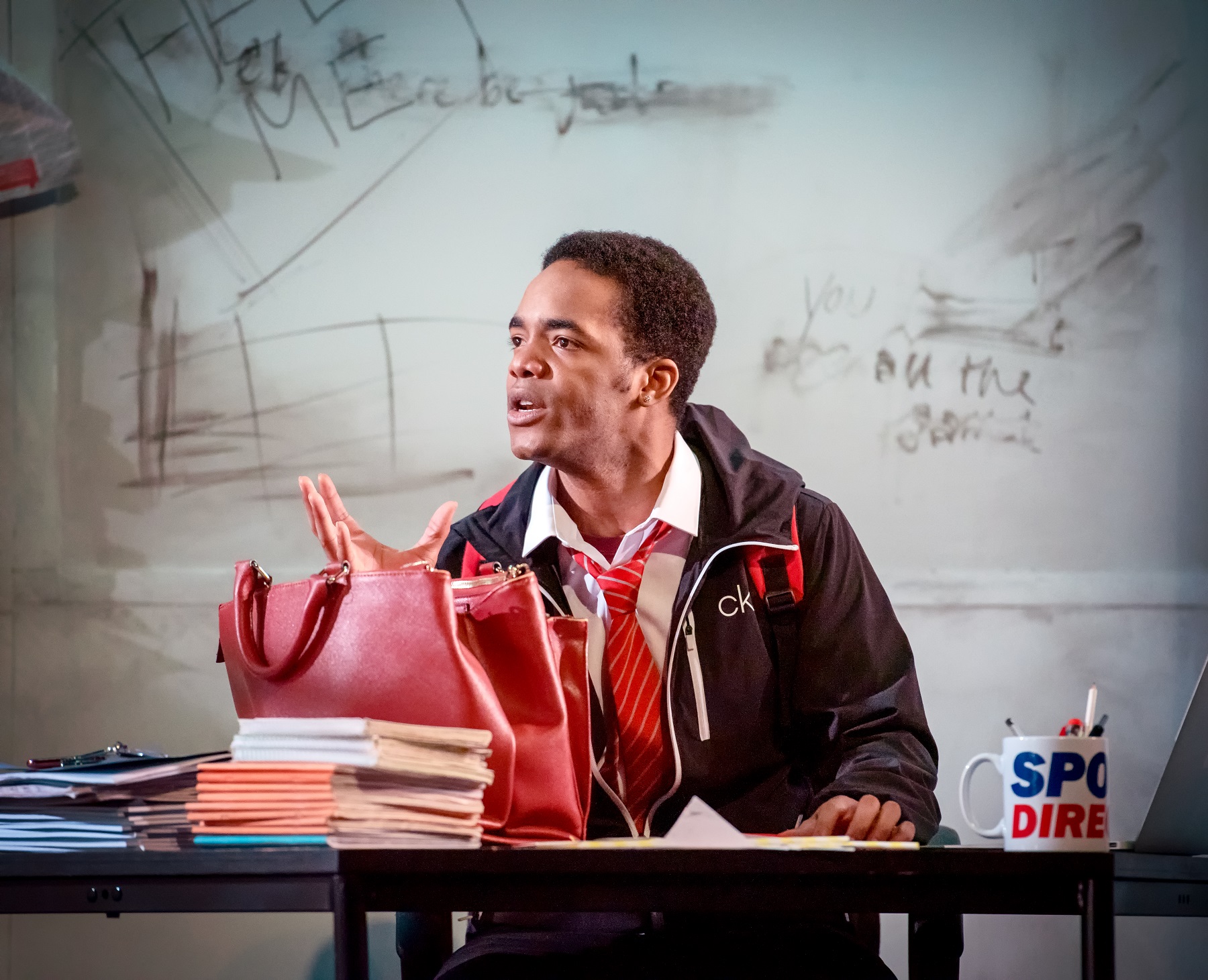 A boy in uniform sits behind a desk speaking and gesticulating to someone off camera