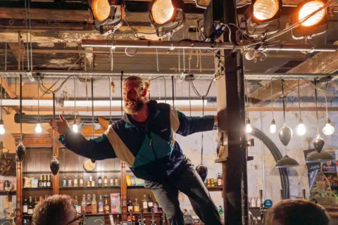 A man stood on a bar, swinging towards an audience from a pillar as he addresses them