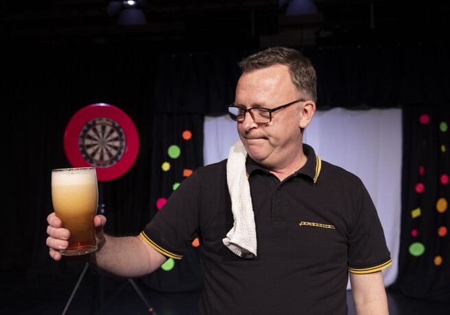 A man holding up a pint of bitter in front of a dartboard