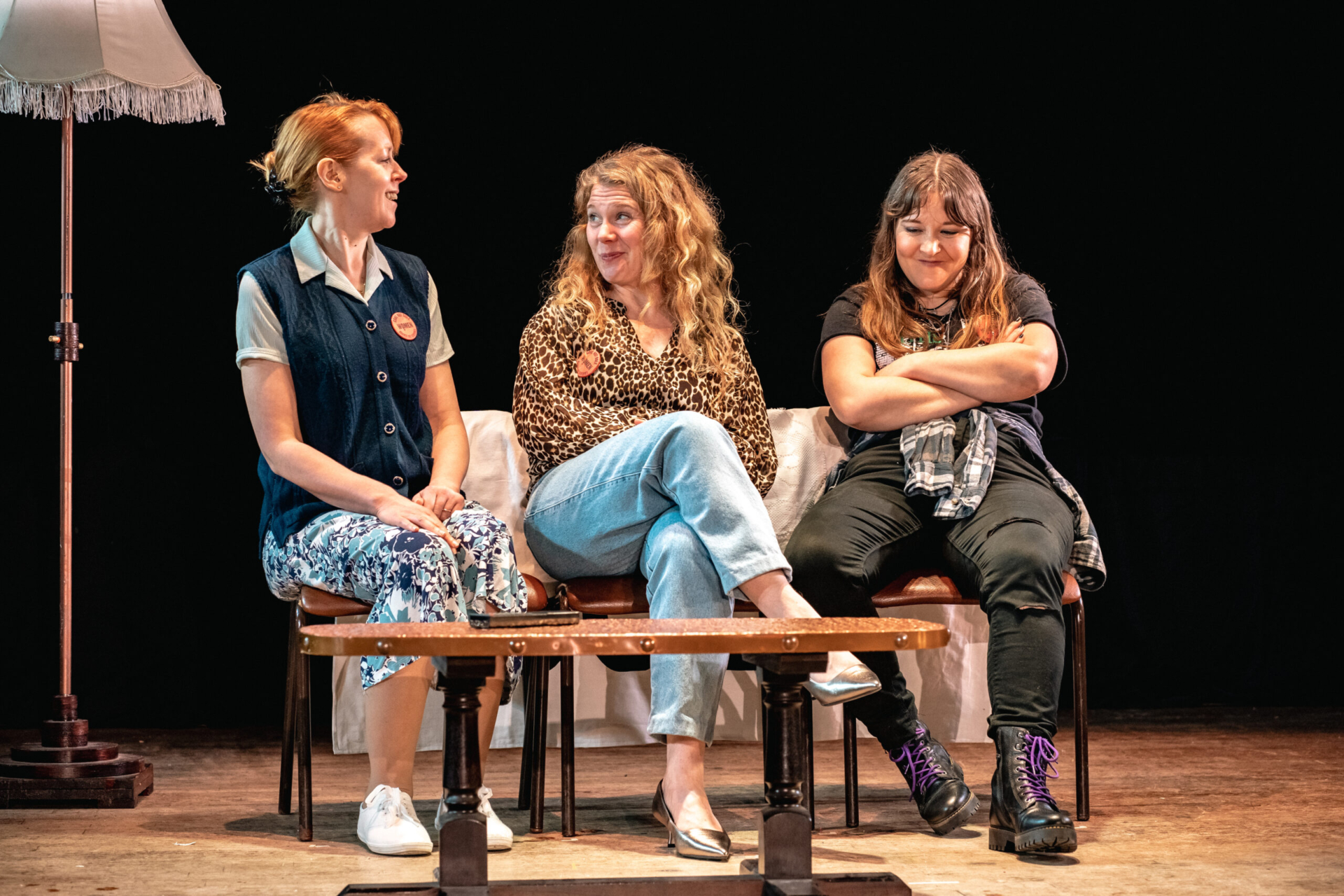 Three women sat on a makeshift sofa behind a coffee table dressed in 80s clothes.