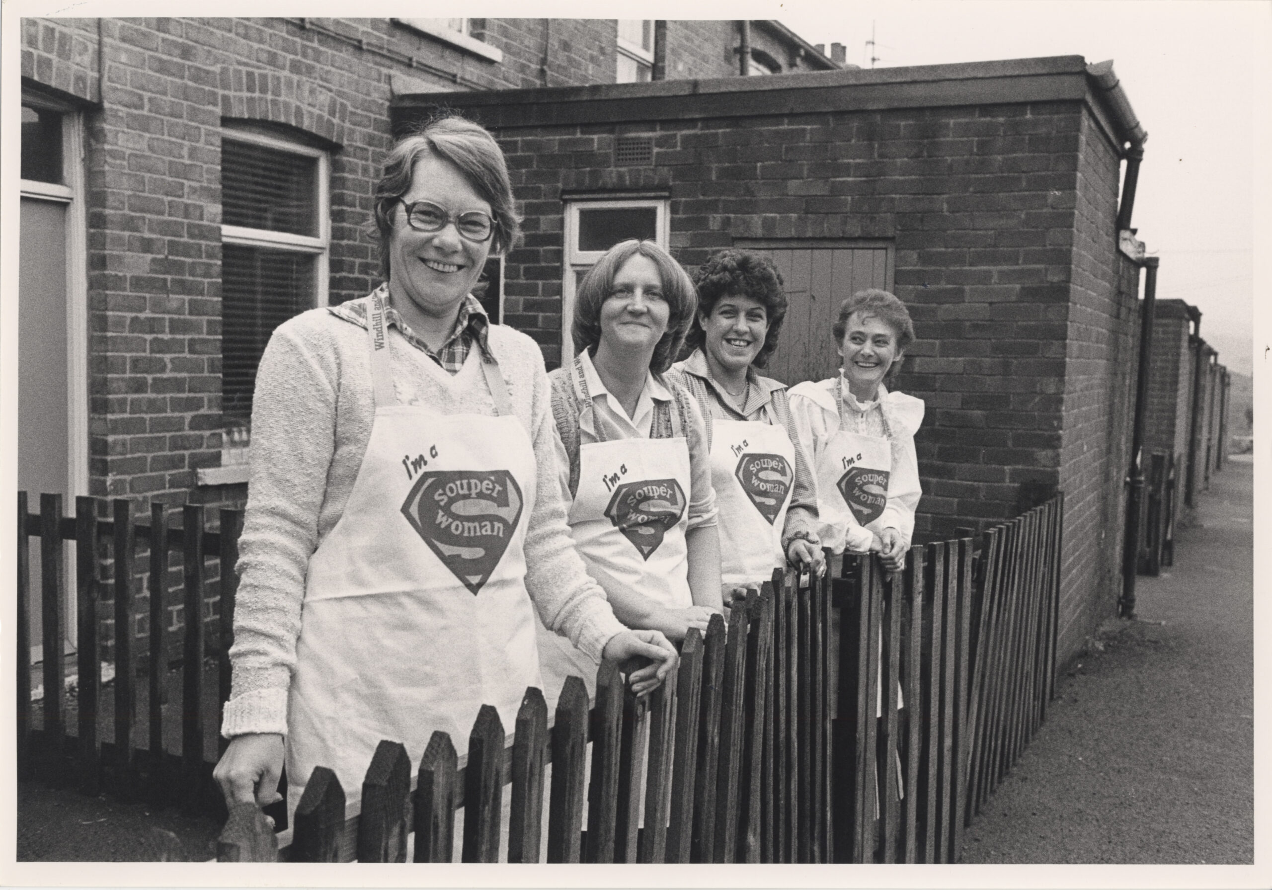Four women dressed in aprons that each read Souper Woman looking over a fence of a back to back in the 80s.
