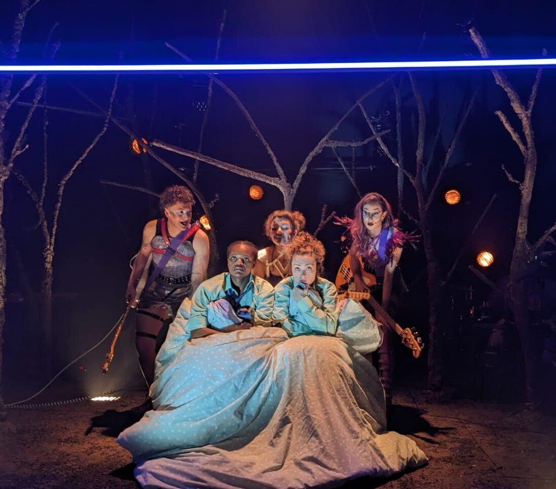 Two women wrapped in a duvet on a stage with a punk band behind them as if watching television in a wood