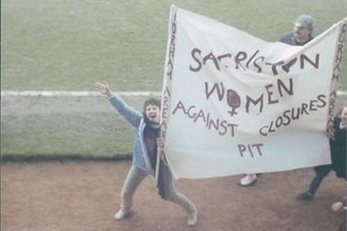A woman holding a white homemade banner that says Sacriston Women Against Pit Closures in red