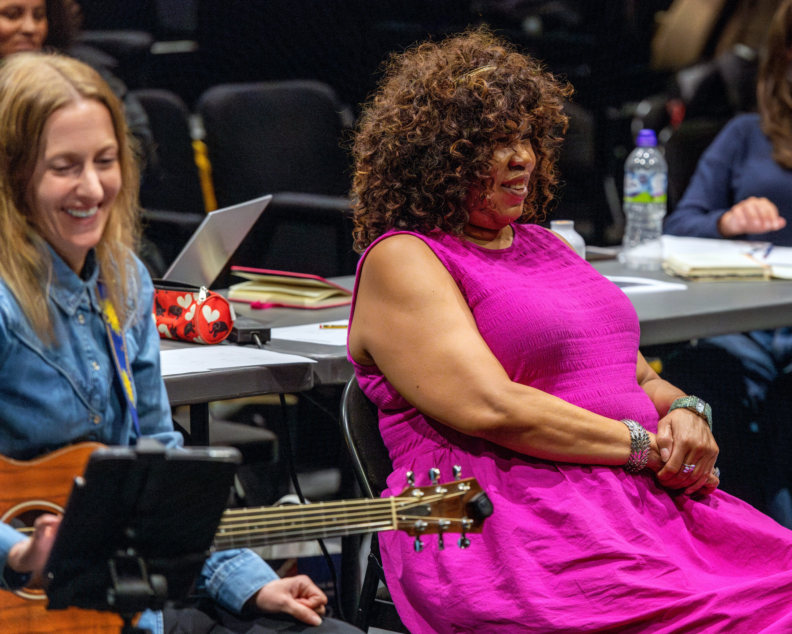 A woman with a guitar sat next to our brilliant artistic director in her pink dress smiling at rehearsals
