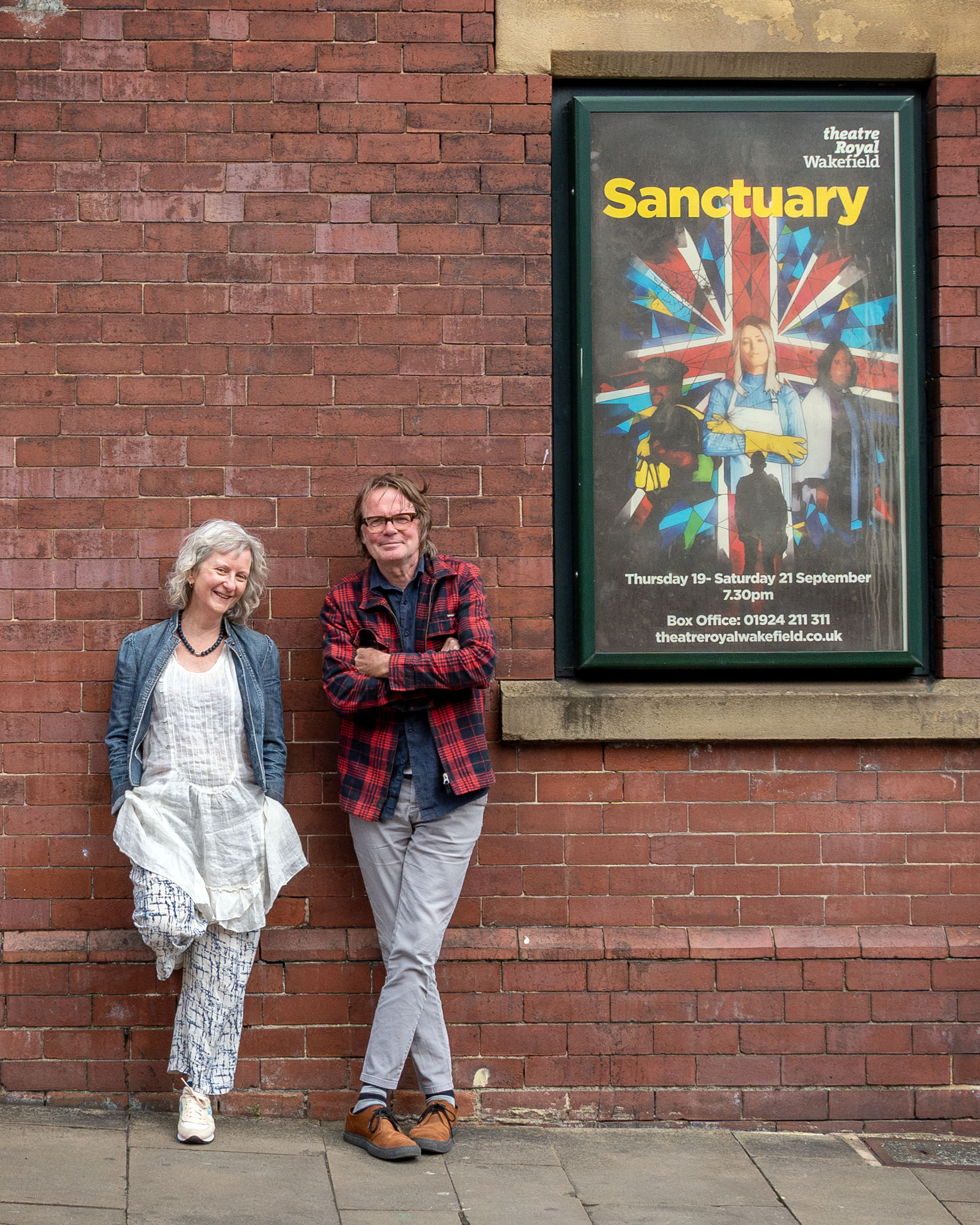 Writers Sarah and Boff leaning against the red brick wall of Theatre Royal Wakefield next to a poster of Sanctuary