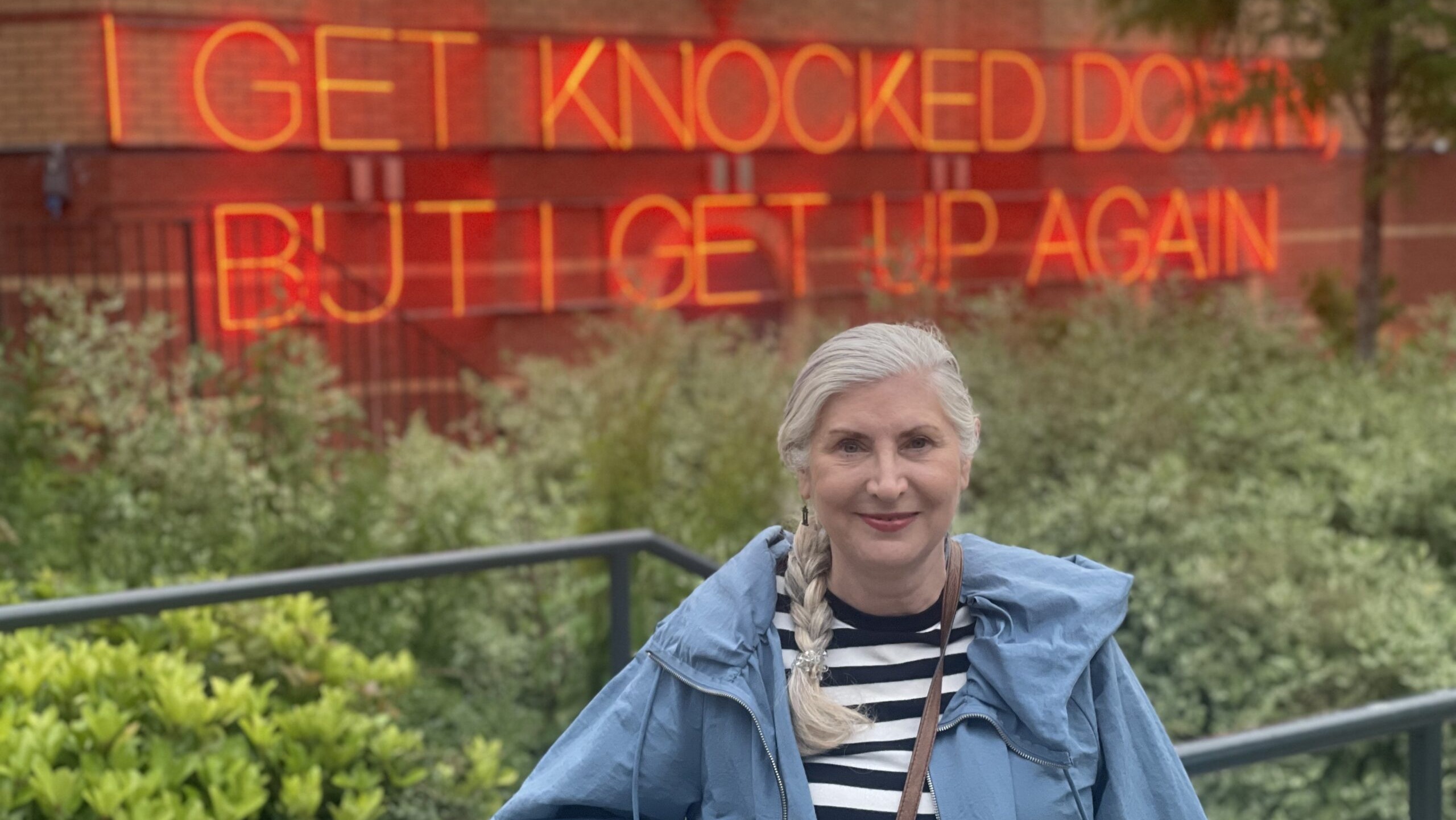 A woman in front of a neon sign that says I get knocked down but I get up again