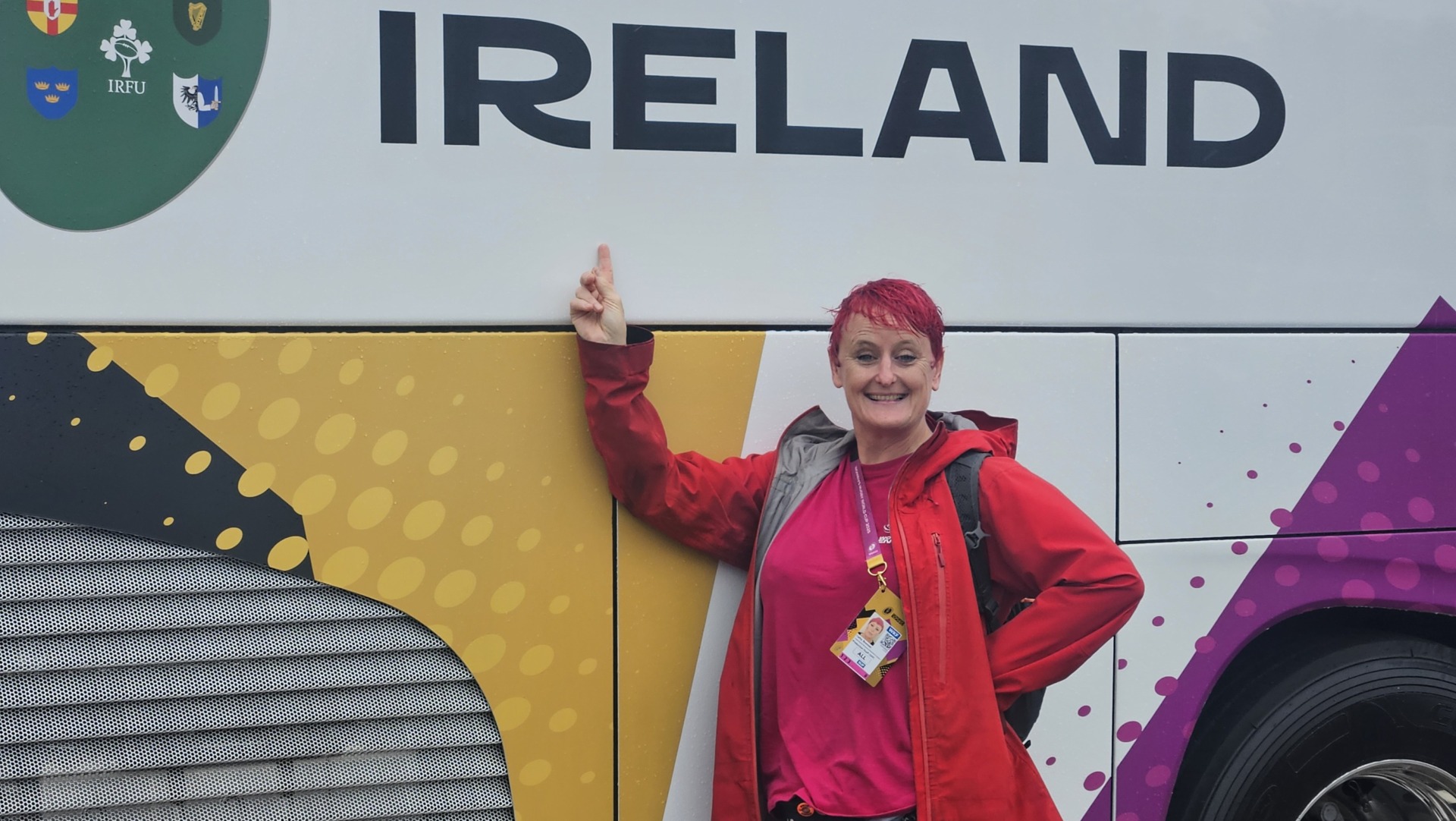 a very wer Cathy smiling next to a coach and pointing to the word Ireland.