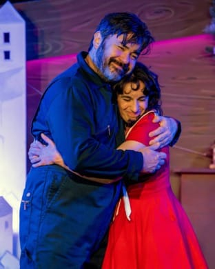 A man in a boiler suit and girl in a red sailor dress hugging
