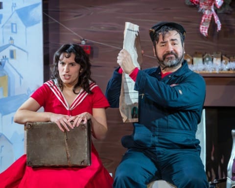 A man in a boiler suit holding up a newspaper between him and a girl with a suitcase on her lap