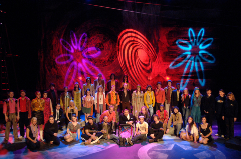 A chorus dressed in 60s and 70s costumes onstage with psychedelic backdrop