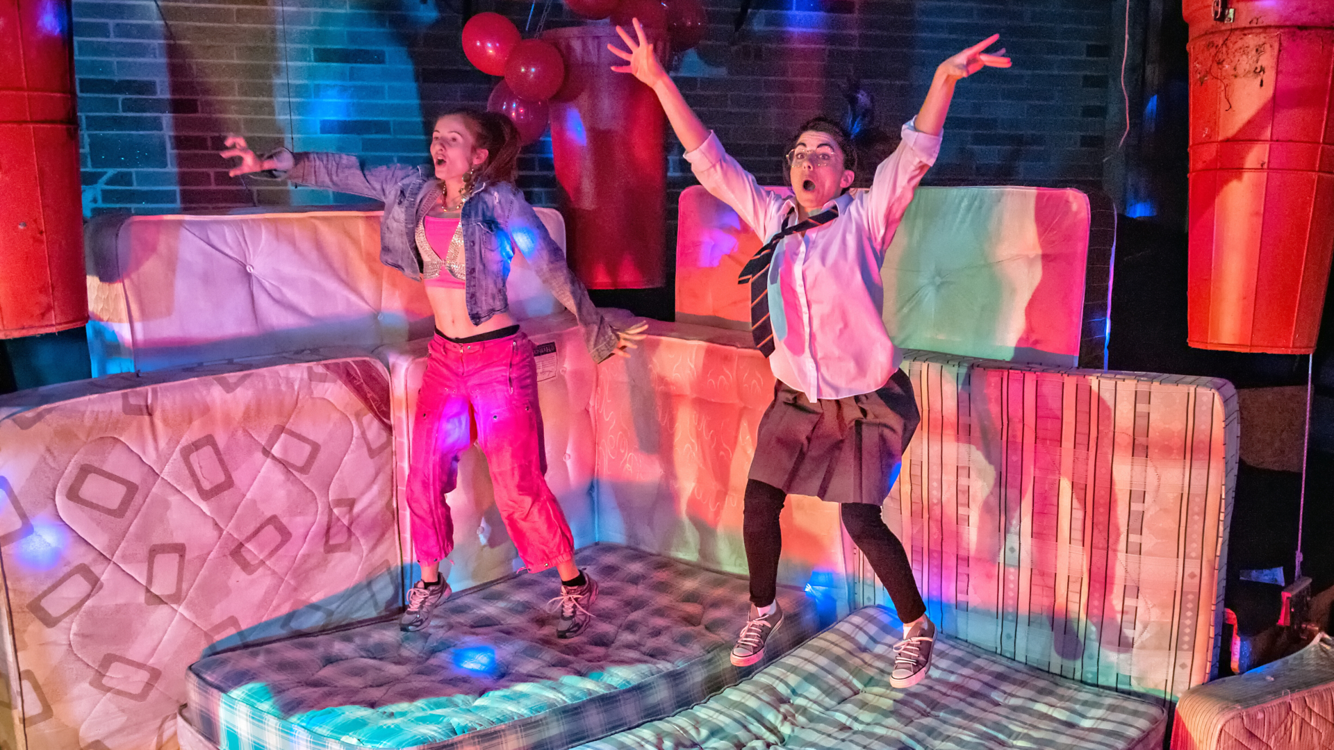 Two young women bouncing on mattresses in a warehouse