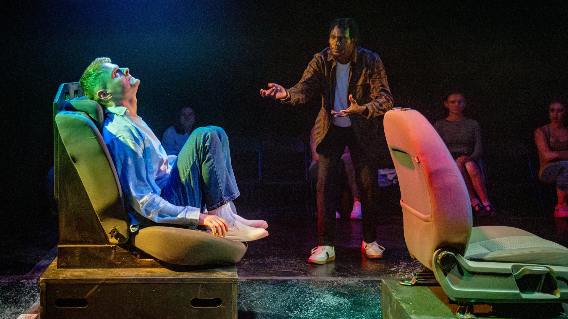 A man sat on a raised car seat stares at the ceiling as a man bargains with him, they are watched by an audience.