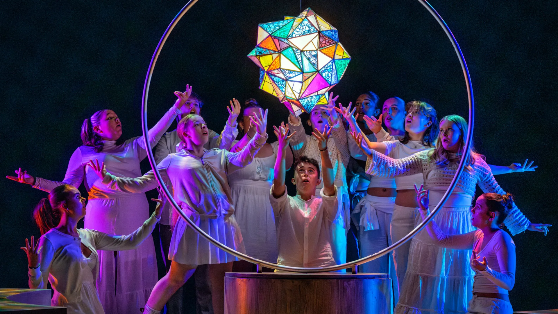 A group of young people in white lifting their hands to a glowing coloured stained glass shape