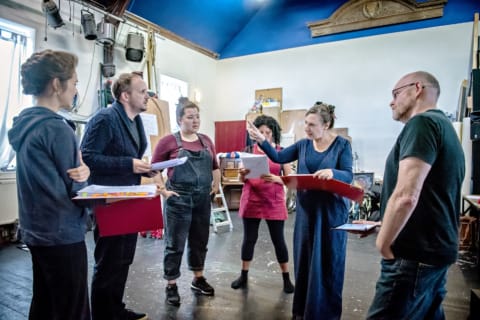 Five actors rehearsing with red folders in hand as the director watches stood closely by
