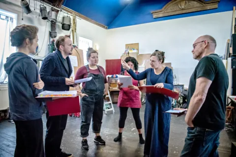 Five actors rehearsing with red folders in hand as the director watches stood closely by