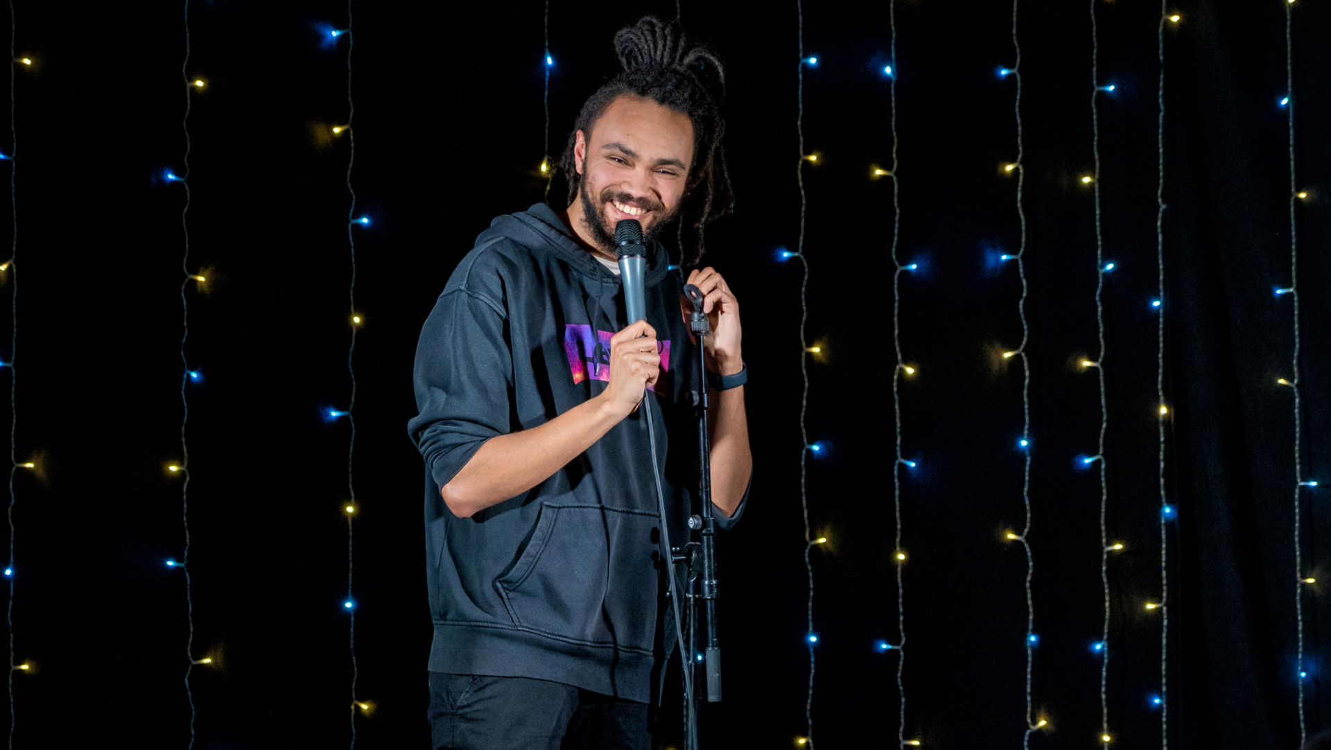 A man holding a microphone and leaning on a mic stand smiling.