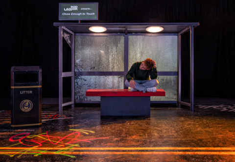 An actor sat crosslegged on stage on a bus shelter bench pre-show