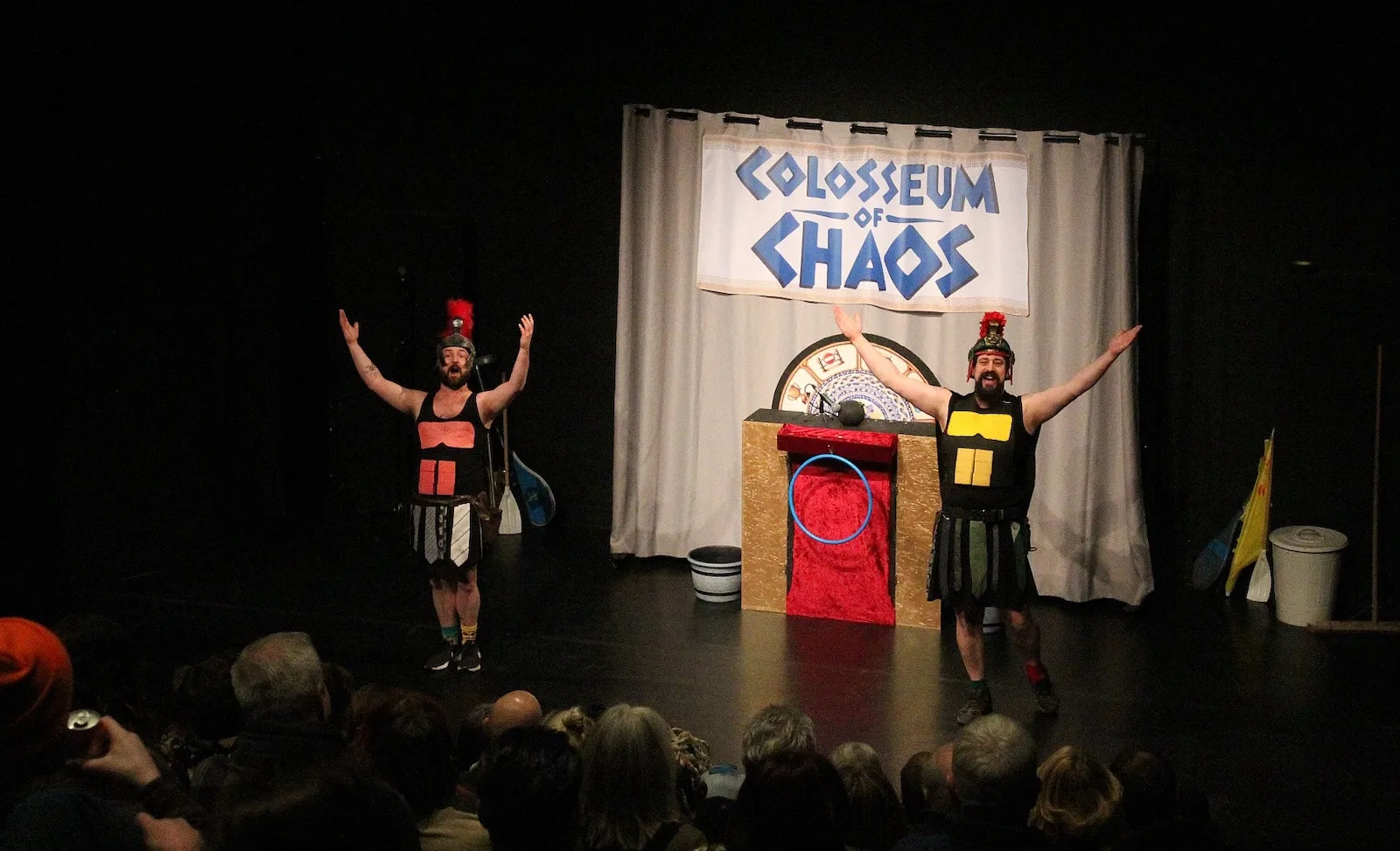 Two gladiators with paper abs stuck on under a sign on stage reading Colloseum of Chaos