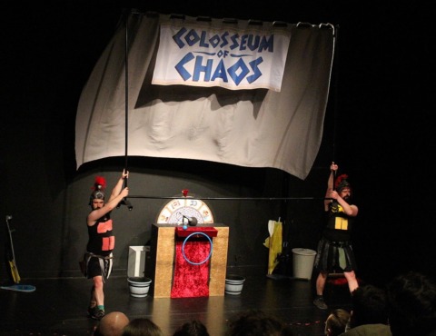 Two gladiators with paper abs stuck on pointing to a huge sign on stage reading Colloseum of Chaos