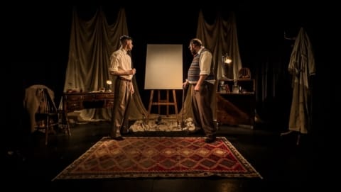 A young and older man stood facing each other in front of a blank canvas on an easel.