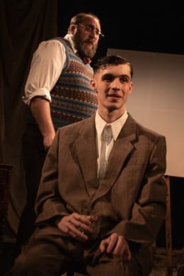 A young man in a suit sat in a chair with an older man looming behind