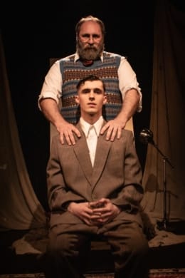 A young man sat in a chair looking uncomfortable as an older man stands behind with hands on his shoulders