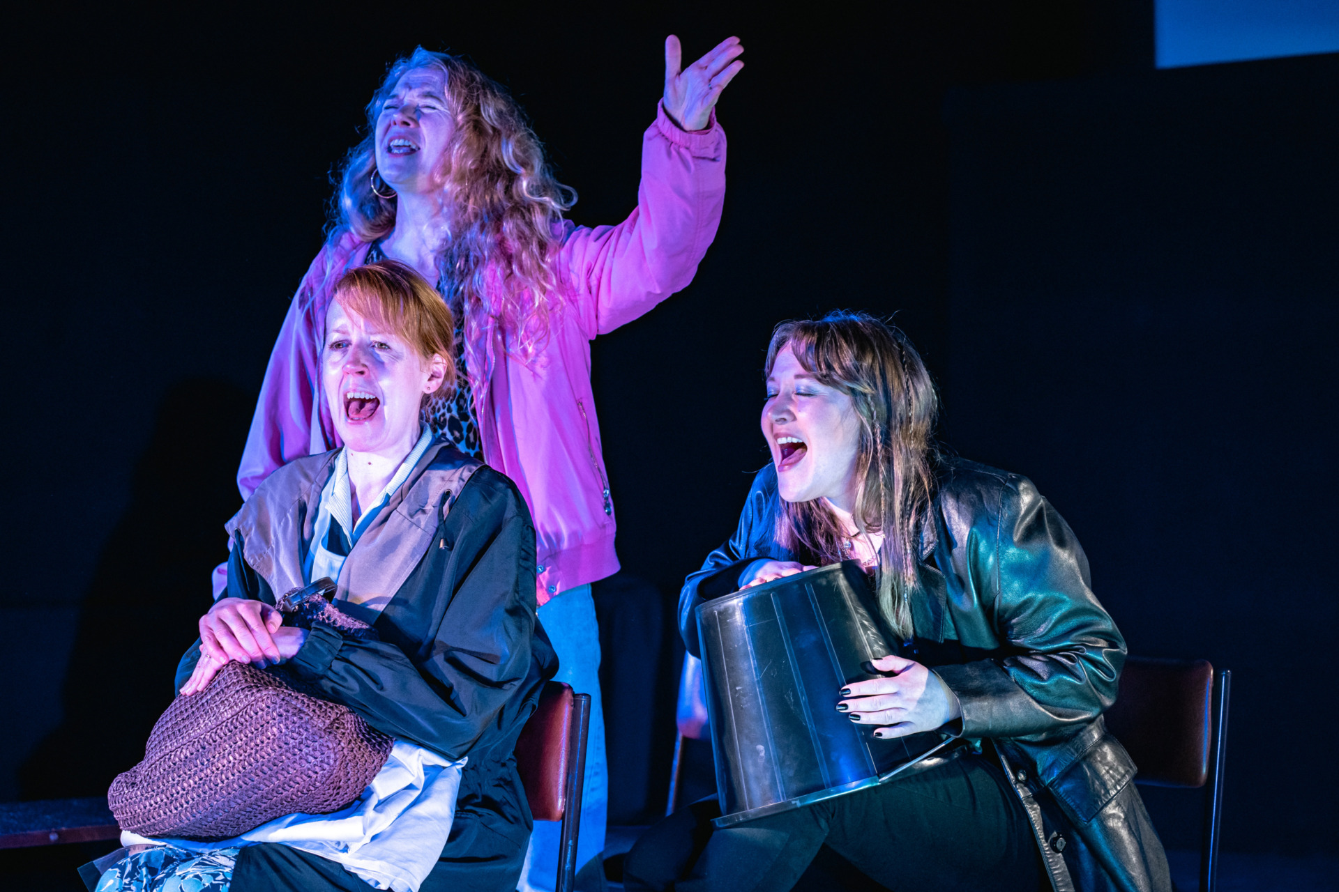 Three women singing as one uses a bucket as a drum