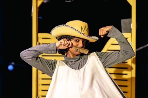 A girl in a stetson holding a comb above her lip like a moustache and flexing her muscles