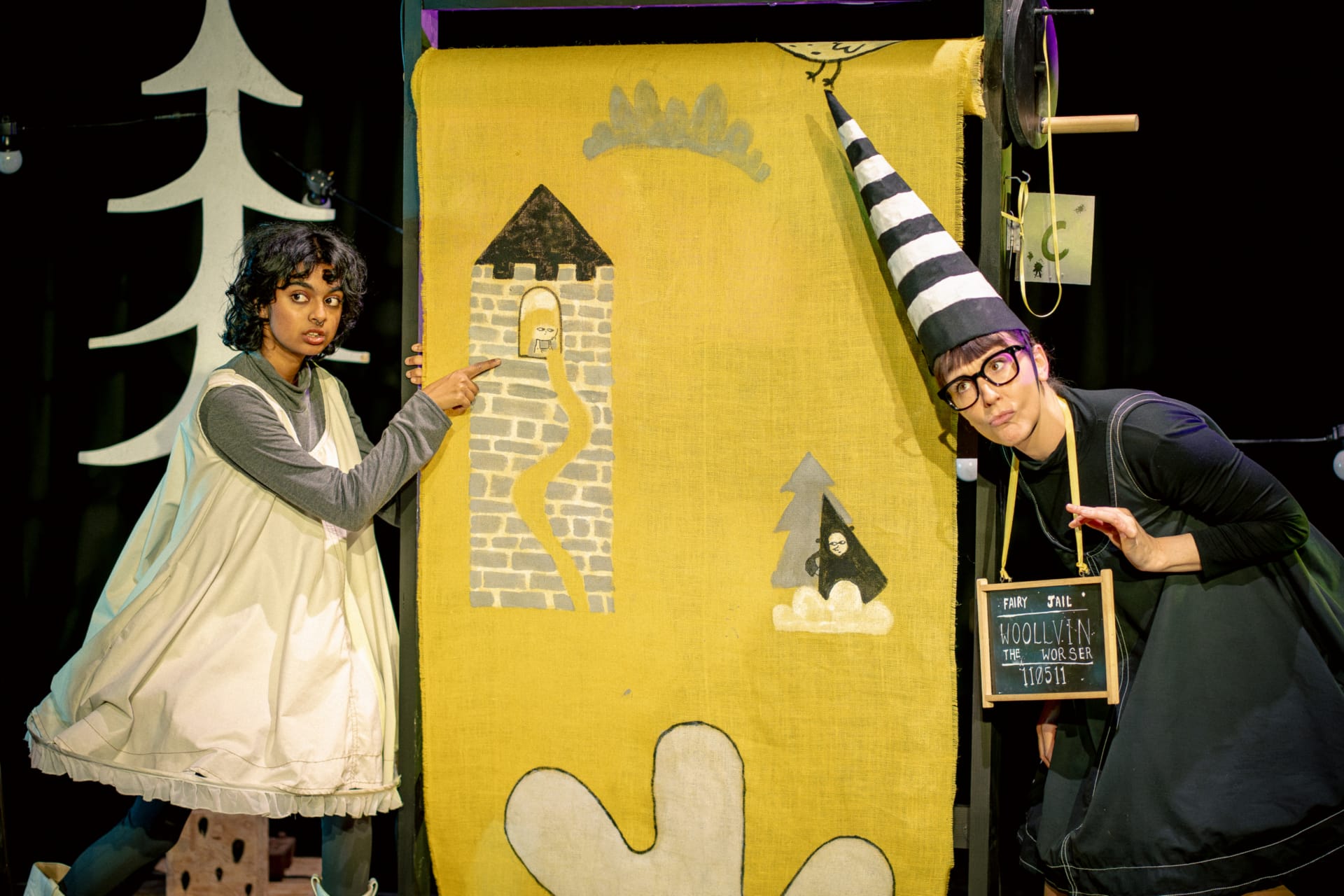 A girl dressed in a pinafore pointing to Rapunzel drawn in her tower with a woman dressed as a dunce peeping round