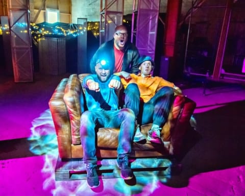 Two men sat on a sofa. One with a helmet torch on and his hands out like driving a vechile. Another man stood behind them.