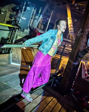 A woman in a denim jacket and purple trousers. She is holding onto a metal bar.