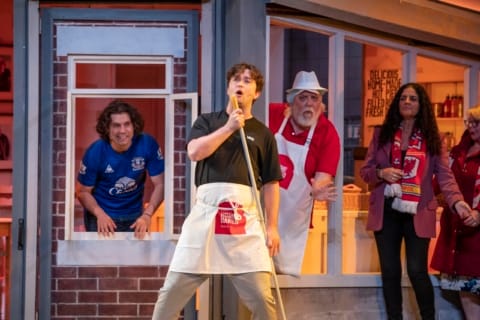 A man using a broom as a microphone. Two people watch on.