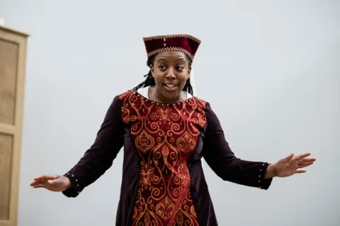 Alicia in a tabard and hat with arms open