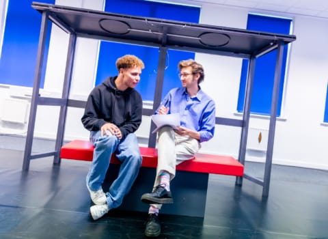 An actor and writer sat on a bus shelter bench in rehearsals discussing a script